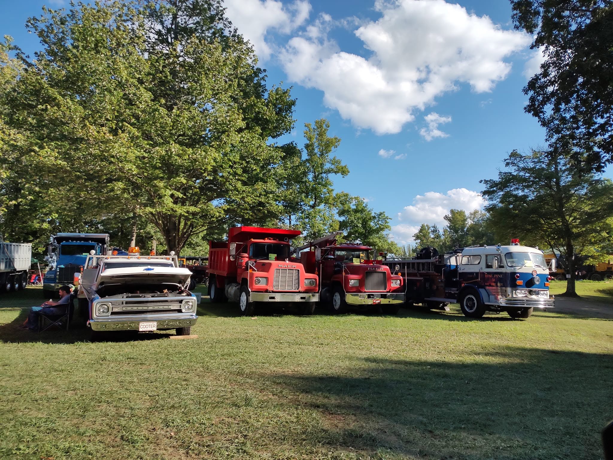 Row of vintage Chevy dump trucks, a red semi, and a vintage bus lined up on the grass
